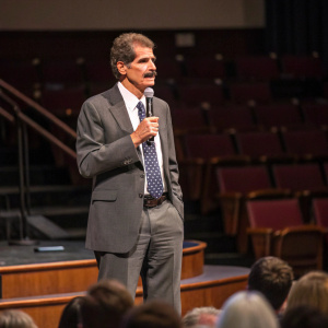 John Stossel, distinguished speaker in a grey suit, white shirt and a dotted tie, addressing a panel. Stage setup, audience in the background.
