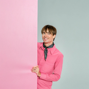 Reeve Carney, Caucasian actor, short brown hair, friendly smile, brown shirt, black bandana. TV Critics Association event backdrop.