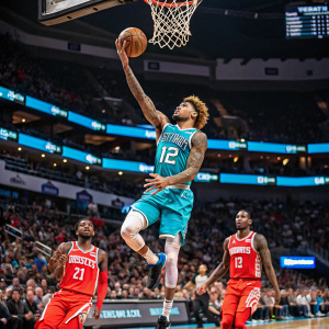 Kelly Oubre Jr., donning Charlotte Hornets jersey number 12, mid shot during intense NBA game. Houston Rockets players scattered around the court. Thrilled fans filling the arena seats.