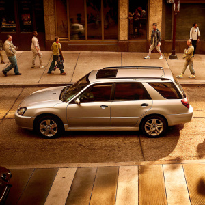 A silver Subaru WRX STI parked on a bustling city street. People in casual attire walk past the car, which is now an SUV model.
