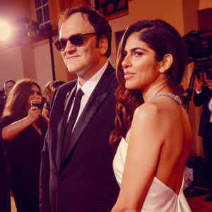Quentin Tarantino and Daniela Tarantino attending the Golden Globe Awards. Quentin in a black suit, sunglasses, standing formally. Daniela in a white evening gown. The backdrop is filled with flashing cameras and celebrities.