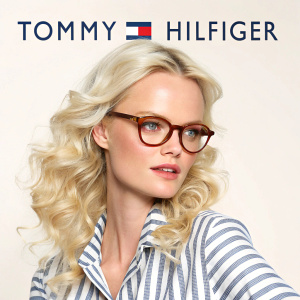 Franziska Knuppe, blonde hair cascading in big curls, stands confidently. Striped shirt, brown glasses. Light background, Tommy Hilfiger logo prominent