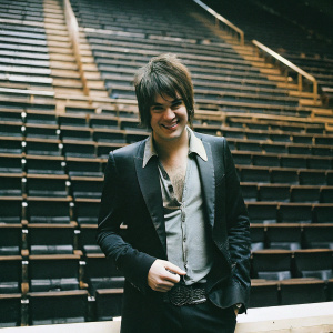 Adam Lambert in a stylish ensemble. Grey shirt, black suit, casual smile. Empty stage backdrop.