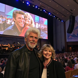 Barry Bostwick and Carol Demas at a musical concert. Large screen showing Grease-themed visuals. Crowd cheering