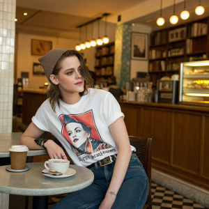 Kristen Stewart, wearing a casual outfit and David Bowie T-shirt, posing in a coffee shop.