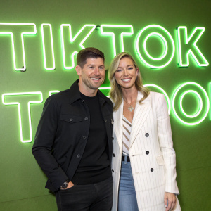 Steven Gerrard and Alex Gerrard, standing in front of a bright neon green wall covered in TikTok logos. Both smiling widely at the camera during a social media influencer event.