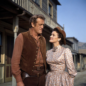 James Arness and Ann Doran on set of *Gunsmoke*. Western-style backdrop. James in a brown vest, Ann in a floral dress.
