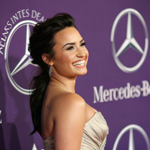 Demi Lovato, smiling widely, at Mercedes-Benz gala. Purple and white logo backdrop