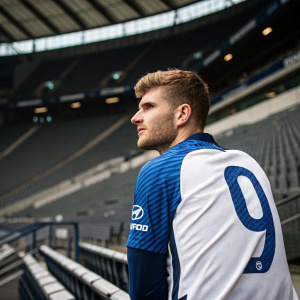 Timo Werner, soccer star, in blue and white jersey with number 9, poses at a press conference. Empty stadium background