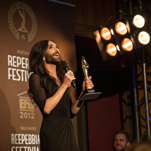 Conchita Wurst presenting an award at Reeperbahn Festival. She is wearing a black dress, illuminated by stage lights. Brown backdrop with logo. Candid shot