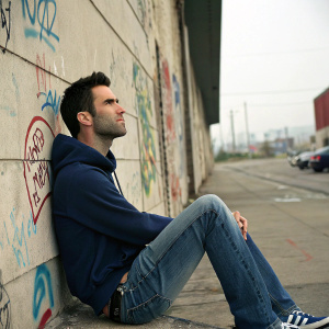 Adam Levine, wearing a navy blue hoodie and faded jeans, sitting against a graffiti-covered concrete wall in a gritty urban setting.