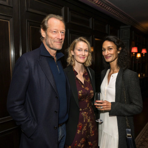Iain Glen, Charlotte Emmerson, and Indira Varma attending a book launch party. Dark wood paneling, dim lighting, elegant attire, casual conversation.