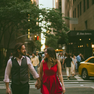 Tamara Ecclestone and Jay Rutland stroll through a busy downtown street. She sports a vibrant red dress. He is in a smart casual outfit. Crowded scene, dynamic movement