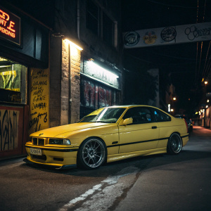 A bright yellow BMW E36 M3, parked in a dimly lit alley. Neon signs and graffiti in the background. Car's exterior, sleek design and lines, in sharp focus