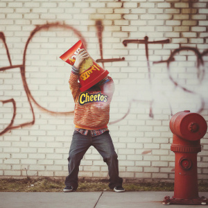 Illustration featuring a vibrant, cartoon character inspired by Frito-Lay's Cheetos mascot. The character is holding a Cheetos bag, positioned energetically in front of a brick wall backdrop with a red fire hydrant. The scene is set to the left of a large graffiti word.