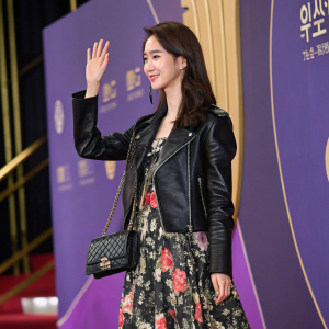 Yoona from Girls' Generation at a glamorous event. Wearing a black leather jacket over a floral dress. Making a wave gesture with a black clutch purse. Vibrant backdrop of purple and gold tones.