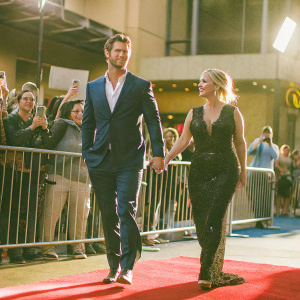 Chris Hemsworth in a sleek navy suit and Christina Applegate in a shimmering black gown at a movie premiere event. Red carpet scene with flashing cameras and excited fans.