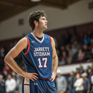 Jarrett Stidham, wearing a blue jersey with number 17, intense expression. Indoor basketball court, spectators blurred in the background.