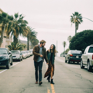 Robert Pattinson and Camilla Belle, laughing and walking down a busy street. Palm trees and parked cars in background.
