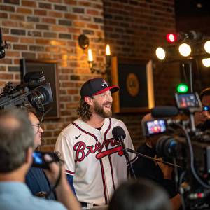 Ian Anderson, in an Atlanta Braves jersey, at a lively press conference. Surrounded by cameras and audio equipment, set against a rustic brick wall backdrop.