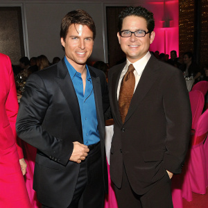 Tom Cruise and J.J. Abrams at a glitzy Hollywood gala. Both exude charm and warmth. Tom in a black suit with blue shirt, J.J. in a dark suit with brown tie. Surrounded by crowd in dimly lit room.