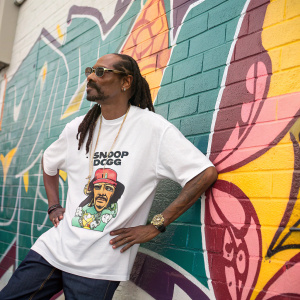 Snoop Dogg, iconic rapper, sporting a unique dreadlock hairstyle. Posing confidently in front of a vibrant graffiti wall. Wears a white t-shirt featuring an illustrated version of himself.