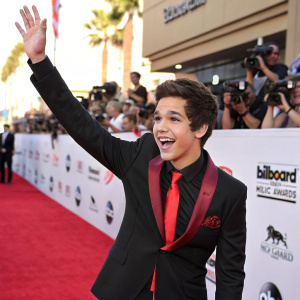 Austin Mahone, a young singer, waving from a red carpet at the Billboard Music Awards. He's wearing a black tuxedo with a red tie.