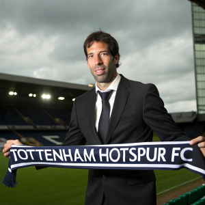 Ruud van Nistelrooy, wearing a black suit and tie, posing for a formal portrait at White Hart Lane stadium. The backdrop features green field and sky. He holds a Tottenham Hotspur FC banner. Lighting is soft, highlighting his facial features and the banner details.
