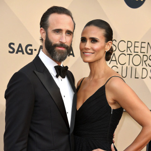 Joseph Fiennes and Maria Dolores Dieguez, dressed formally, at the Screen Actors Guild Awards. Fiennes in a tuxedo, Dieguez in an elegant black gown. They pose confidently against a neutral backdrop.