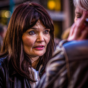 Helena Christensen, with deep brown eyes and straight dark hair, wearing a casual outfit including a black leather jacket, captured in a candid street photography style. Background blurred, focusing solely on her expression and features.