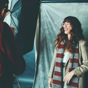 Olivia Thirlby, captured in a candid moment. Long wavy brown hair, wide smile, beige plaid blouse, red and gray knitted scarf. Gray studio backdrop, soft, natural lighting