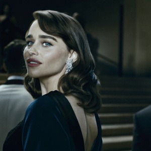 Close-up of Emilia Clarke, fair-skinned actress, with shoulder-length brown hair and striking makeup. She's in a dark, fitted dress and large silver earrings. Blurred background at a glamorous event.