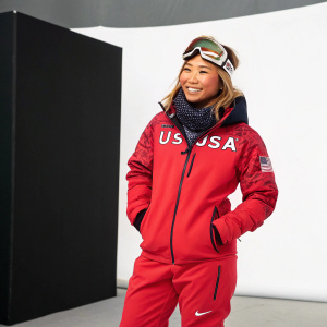 Chloe Kim, Asian female Olympian, posing in a Nike tracksuit with USA emblem. Standing in front of a minimalist black backdrop. Friendly smile, wearing goggles and a scarf.
