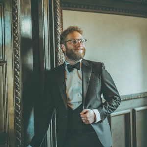 Seth Rogen in a sleek black tuxedo, with a beard and glasses, posing at the Sundance Film Festival. Black background