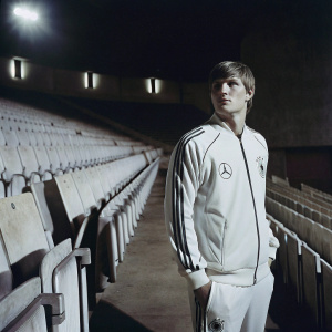 Toni Kroos, dressed in Adidas and Emirates gear, standing in an empty stadium. The seats are filled with empty chairs, lights cast shadows in the dimly lit arena