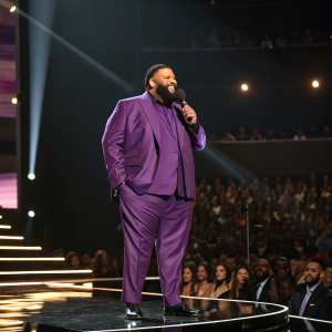 DJ Khaled, with a beard, in a purple suit, center stage at the Billboard Music Awards. Spotlight shining, audience seated