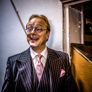 Edward Hibbert, actor, dressed in a black pinstripe suit, white shirt and pink tie, standing in front of a plain white wall. Cheerful expression. Event promotion for Broadway show 'The Drowsy Chaperone'.
