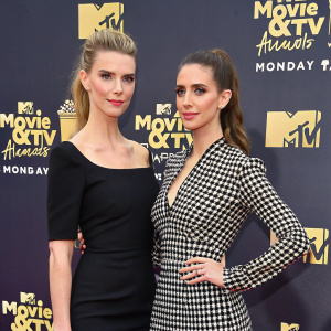 Betty Gilpin and Alison Brie on the MTV Movie & TV Awards red carpet. Betty in a simple black dress, Alison in a checkered blouse. Dark backdrop with MTV logos