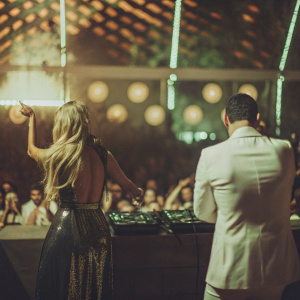 Paris Hilton and AfroJack at an indoor concert. Paris in a shimmering black dress. AfroJack in a white suit. Crowd in the background