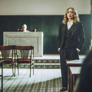Elisabeth Röhm as ADA Serena Southerlyn. Dressed in a sharp, black courtroom suit. Standing confidently in a dimly lit, empty courtroom.