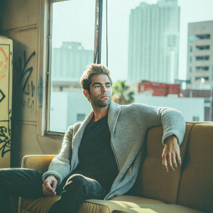 Chace Crawford, wearing a gray sweater and black t-shirt, posing for a casual photo shoot. Brightly lit, modern urban setting with graffiti and cityscape in the background.