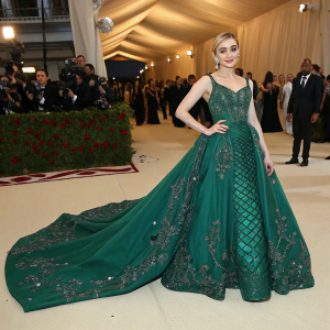 Meg Donnelly at the Met Gala. Wearing an elegant, floor-length emerald green dress with intricate beadwork and long train. Poses in front of a crowded event space.