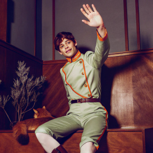 3D render of Gon Freecss from Hunter x Hunter. Wearing green outfit with orange accents, waving hand. Playful pose against dark grey backdrop with small plants and a brown squirrel-like creature nearby.