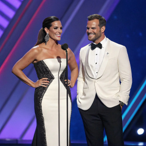 Gaby Espino and Mark Tacher at the 2019 Billboard Latin Music Awards, elegantly dressed. Gaby in a sophisticated white and black gown, Mark in a formal tuxedo. They stand on a stage with a blue and purple backdrop.