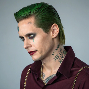 Jared Leto as the Joker. Wearing a dark green hair and gothic makeup. Piercings and tattoos visible. Dark maroon shirt and chain necklace. Studio shot.