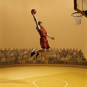 Mac McClung in vibrant red jersey leaping for a dunk. Audience buzzing in the background. Basketball court illuminated under bright lights.