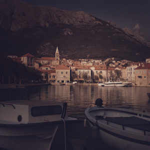 Makarska, Croatia: A scenic coastal town featuring colorful houses and medieval structures lining the hillsides. Calm azure waters mirror the buildings and create a serene atmosphere. Several fishing boats are docked along the shore.