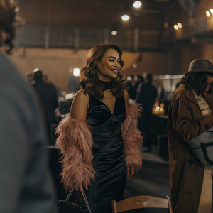 Preity Zinta, wearing a black dress with a pink fur stole, smiling warmly at a bustling indoor event.
