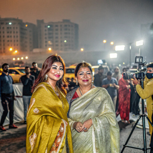 Ayesha Takia Azmi and her mother Farida Takia at a bustling Bollywood premiere. Both in vibrant, traditional sarees. City lights and paparazzi in background.
