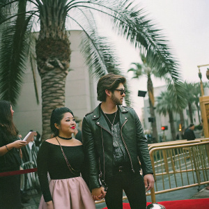 Cierra Ramirez and Jeff Wittek attending a Hollywood film premiere. Cierra in a black blouse and pink skirt, adorned with jewelry. Jeff in a black graphic tee and leather jacket. Palm trees and a golden barrier visible in the background.